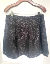 Load image into Gallery viewer, A black lace mini skirt with a subtle floral damask pattern, hanging on a wooden hanger against a plain white wall.