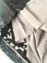 Load image into Gallery viewer, Close-up of a light beige or taupe pleated fabric garment, likely a skirt or dress section, layered over a black and white patterned textile.