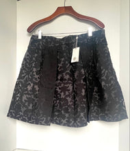 Load image into Gallery viewer, A black and gray floral jacquard pleated mini skirt hanging on a wooden hanger against a white wall, with a white product tag attached.