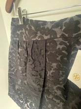 Load image into Gallery viewer, A close-up of a pleated skirt with a dark gray and black jacquard pattern, hanging on a metal hanger with a white paper tag attached.