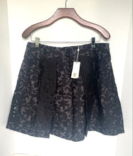 Load image into Gallery viewer, A black and gray floral jacquard pleated skirt hanging on a wooden hanger against a white background, with a white paper tag attached.
