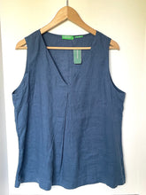 Load image into Gallery viewer, A sleeveless slate blue linen top hanging on a wooden hanger against a plain off-white wall. The top features a V-neckline and side pockets, with a green brand tag visible.