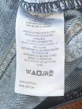 Load image into Gallery viewer, Close-up of a white garment care label sewn onto blue-gray denim fabric, showing care instructions in English and French.
