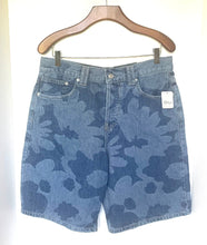 Load image into Gallery viewer, A pair of blue denim shorts with a dark blue abstract floral or cloud-like pattern, hanging on a wooden hanger against a plain white wall. A white brand tag is visible on the right pocket.