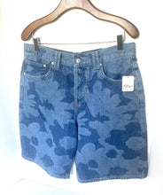 Load image into Gallery viewer, Light blue denim shorts with a darker blue camouflage pattern, hanging on a wooden hanger against a white wall. A white tag is attached to the right pocket.