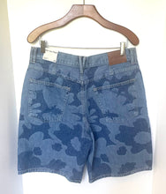 Load image into Gallery viewer, Blue camouflage denim shorts hanging from a wooden hanger against a white background, showing the back pockets and waistband.