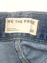 Load image into Gallery viewer, Close-up of a denim waistband showing a cream-colored fabric brand label that reads 'WE THE FREE' with size '28' and 'MADE IN TURKEY' printed on it.