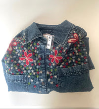 Load image into Gallery viewer, A folded denim shirt jacket with colorful floral embroidery and applique on the front, shown on a plain light background.