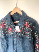 Load image into Gallery viewer, A blue denim button-up shirt jacket with colorful embroidered floral patterns on the chest and sleeves, hanging on a wooden hanger against a plain off-white wall.