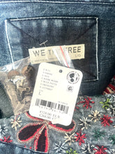 Load image into Gallery viewer, Close-up of a denim garment showing the back pocket, a sewn-on brand label reading WE THE FREE, a hanging product tag with barcode and size info, and decorative embroidered floral details in red, green, and white on the fabric.