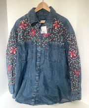 Load image into Gallery viewer, A blue denim button-up shirt jacket with red and pink floral embroidery and a small multicolor floral print on the chest and sleeves, hanging on a wooden hanger against a white wall.