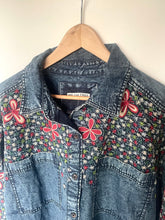 Load image into Gallery viewer, A denim button-up shirt jacket with colorful embroidered butterflies and floral patterns, hanging on a wooden hanger against a plain light background.
