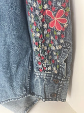 Load image into Gallery viewer, Close-up of folded blue denim jeans showing a decorative embroidered floral panel on the leg with a large red flower and smaller multicolored flowers.