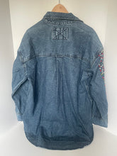 Load image into Gallery viewer, A blue denim jacket shown from the back, featuring a patch on the upper left with an X symbol and a small floral embroidered detail on the upper right shoulder, displayed on a mannequin torso against a plain white background.