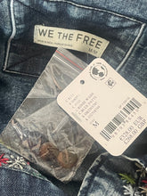 Load image into Gallery viewer, Close-up of a pair of dark blue denim jeans showing the back pocket with a sewn-on 'WE THE FREE' label, a white retail price tag, and a small clear plastic bag containing buttons.