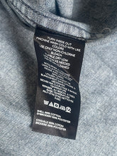 Load image into Gallery viewer, A close-up of a black garment care label sewn onto the inside collar of a gray denim or twill fabric garment, showing white printed washing and care instructions.