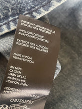Load image into Gallery viewer, A close-up of a dark brown garment care label sewn onto blue-gray denim fabric, showing printed text including material composition, country of origin, and manufacturer details.
