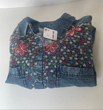 Load image into Gallery viewer, A folded pair of blue denim jeans with colorful embroidered floral patterns and red bow details, featuring a white retail tag attached, photographed on a plain white background.