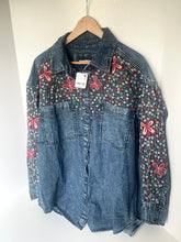 Load image into Gallery viewer, A blue denim button-up shirt jacket hanging on a wooden hanger against a white wall. The shirt features a floral embroidered yoke across the chest and upper sleeves, with pink and red flower motifs over a small multicolor floral pattern.