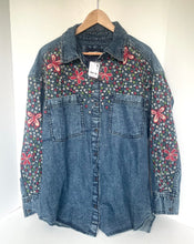 Load image into Gallery viewer, A blue denim button-up shirt jacket with floral embroidery on the chest and sleeves, hanging on a wooden hanger against a white background.