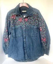 Load image into Gallery viewer, A blue denim button-up shirt jacket with red embroidered floral patterns and small multicolor floral details across the chest and sleeves, hanging on a wooden hanger against a plain light background.