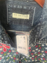 Load image into Gallery viewer, Close-up of a dark blue denim garment showing the back pocket with a cream 'WE THE FREE' label and a hanging price tag, next to a floral-patterned textile.