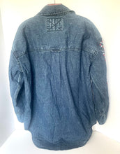 Load image into Gallery viewer, A blue denim jacket shown from the back, hanging against a plain white background. The back features a large patch pocket with a stitched X design, long sleeves, and a small embroidered patch on the left sleeve.