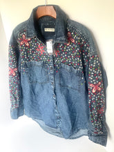 Load image into Gallery viewer, A blue denim button-up shirt jacket with colorful floral embroidery on the chest and sleeves, hanging on a wooden hanger against a plain white wall.
