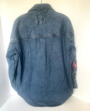 Load image into Gallery viewer, A blue denim jacket shown from the back, hanging against a plain white background. The back features a central seam, a decorative stitched patch on the upper back, and colorful embroidered details on the right sleeve.
