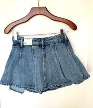 Load image into Gallery viewer, A light blue denim mini skirt with a pleated front and raw hems, hanging from a wooden hanger against a plain off-white wall. The brand label reads 'The One Store'.