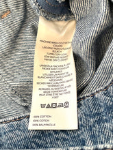 Load image into Gallery viewer, Close-up of a white garment care label sewn onto blue denim fabric, showing washing instructions and cotton content.