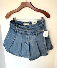 Load image into Gallery viewer, A pair of blue denim pleated shorts hanging from a wooden hanger against a plain white wall, with a visible brand label and a product tag attached.