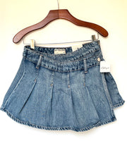 Load image into Gallery viewer, A light blue denim pleated mini skirt hanging from a wooden hanger against a plain off-white wall. The brand label 'GAY THE GIRL' is visible inside the waistband, and a white retail tag is attached to the right side.