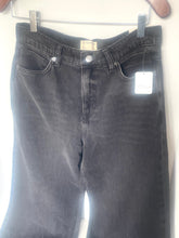 Load image into Gallery viewer, A pair of dark gray washed denim jeans hanging on a metal hanger against a white wall, with a white retail tag attached to the right side.