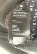 Load image into Gallery viewer, Close-up of a garment's interior label showing the brand 'WE THE FREE' and fabric composition details, with dark gray fabric visible around it.