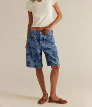Load image into Gallery viewer, A model wearing blue camouflage-patterned denim shorts, a white cropped t-shirt, and brown woven flat shoes, standing against a plain beige studio background.