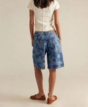 Load image into Gallery viewer, A model shown from the back wearing blue camouflage-patterned denim shorts, a white short-sleeve button-up shirt, and brown flat sandals, set against a plain beige studio background.