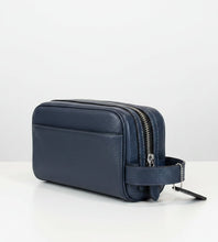 Load image into Gallery viewer, A dark blue pebbled leather zippered pouch or cosmetic case with a matching leather strap and a hanging tag, placed on a light wood surface against a plain light gray wall.