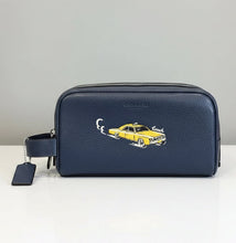 Load image into Gallery viewer, A navy blue pebbled leather Coach zip-around pouch with a wristlet strap and a yellow taxi graphic on the front, displayed on a light wood surface against a neutral gray background.