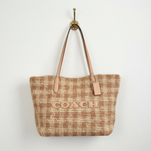 Load image into Gallery viewer, A Coach woven straw tote bag in tan and beige, displayed on a cream-colored mannequin torso. The bag features tan leather shoulder straps, a matching leather hangtag, and the brand name 'COACH' embossed on the front.
