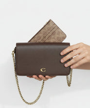 Load image into Gallery viewer, A woman holds a brown pebbled leather crossbody bag with a gold chain strap and a gold logo clasp. A matching beige and brown patterned zip pouch is tucked behind the bag in a blurred boutique setting.