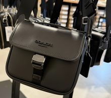 Load image into Gallery viewer, Black Coach handbag held in a store setting with other bags in the background.