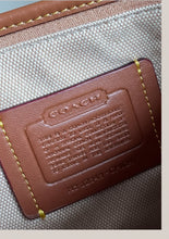 Load image into Gallery viewer, Close-up of a Coach accessory showing a brown leather embossed logo patch with text and a beige woven fabric background.