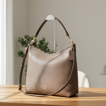Load image into Gallery viewer, Brown leather handbag with gold accents on a light surface with a plant and books in the background