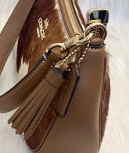 Load image into Gallery viewer, A close-up of a brown and brown-foam shoulder bag with gold-tone hardware, a tassel charm, and a Coach logo on the leather strap.