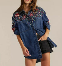 Load image into Gallery viewer, A model wears a blue denim shirt dress featuring colorful floral embroidery on the yoke and sleeves, styled with black denim shorts against a solid beige background.