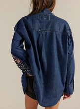 Load image into Gallery viewer, A woman wearing an oversized dark blue denim shirt with colorful floral embroidery on the sleeves, shown from the back against a plain beige background.