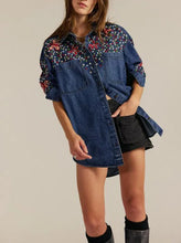 Load image into Gallery viewer, A model wears an oversized blue denim shirt dress featuring colorful floral embroidery on the chest and sleeves, styled with a black denim mini skirt and black boots against a plain beige background.