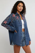 Load image into Gallery viewer, A woman wearing an oversized blue denim button-up shirt with colorful floral embroidery on the chest and sleeves, paired with denim shorts against a plain light gray background.