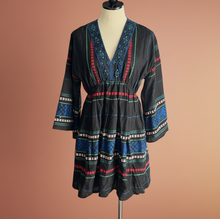 Load image into Gallery viewer, A black long-sleeve dress with colorful geometric patterns in blue, red, green, and white, displayed on a mannequin in a home interior setting.